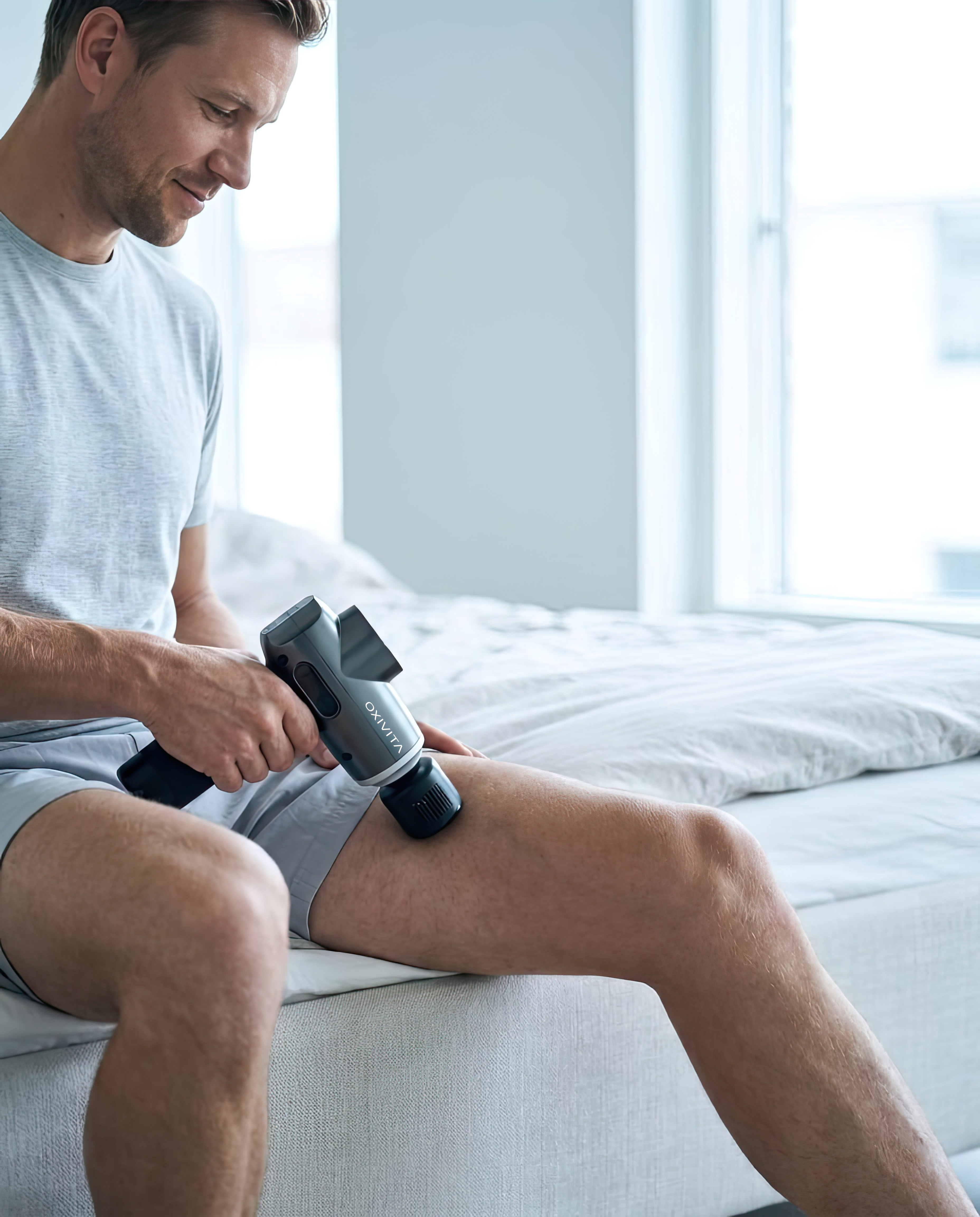 Man masserar låret med Oxivita Core M1 massagepistol med Duo Head med värme- och kylfunktion, sittande på sängen hemma
